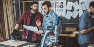 Two men in plaid shirts discussing piece of paper in construction workshop.