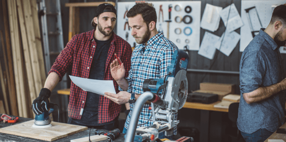 Two men in plaid shirts discussing piece of paper in construction workshop.