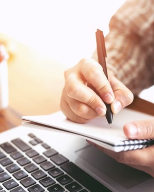 someone writing in a notebook with a laptop in front of them