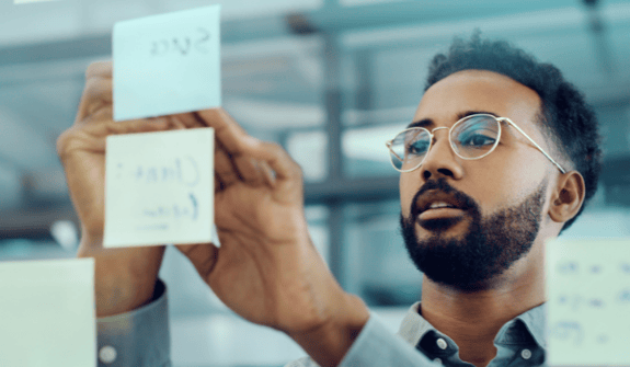 man wearing glasses writing on sticky notes on glass wall