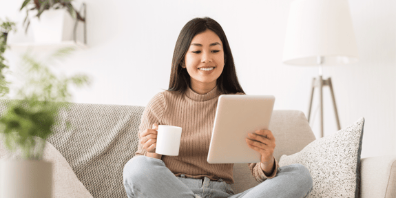 smiling woman reading from an e-reader on her couch with a mug